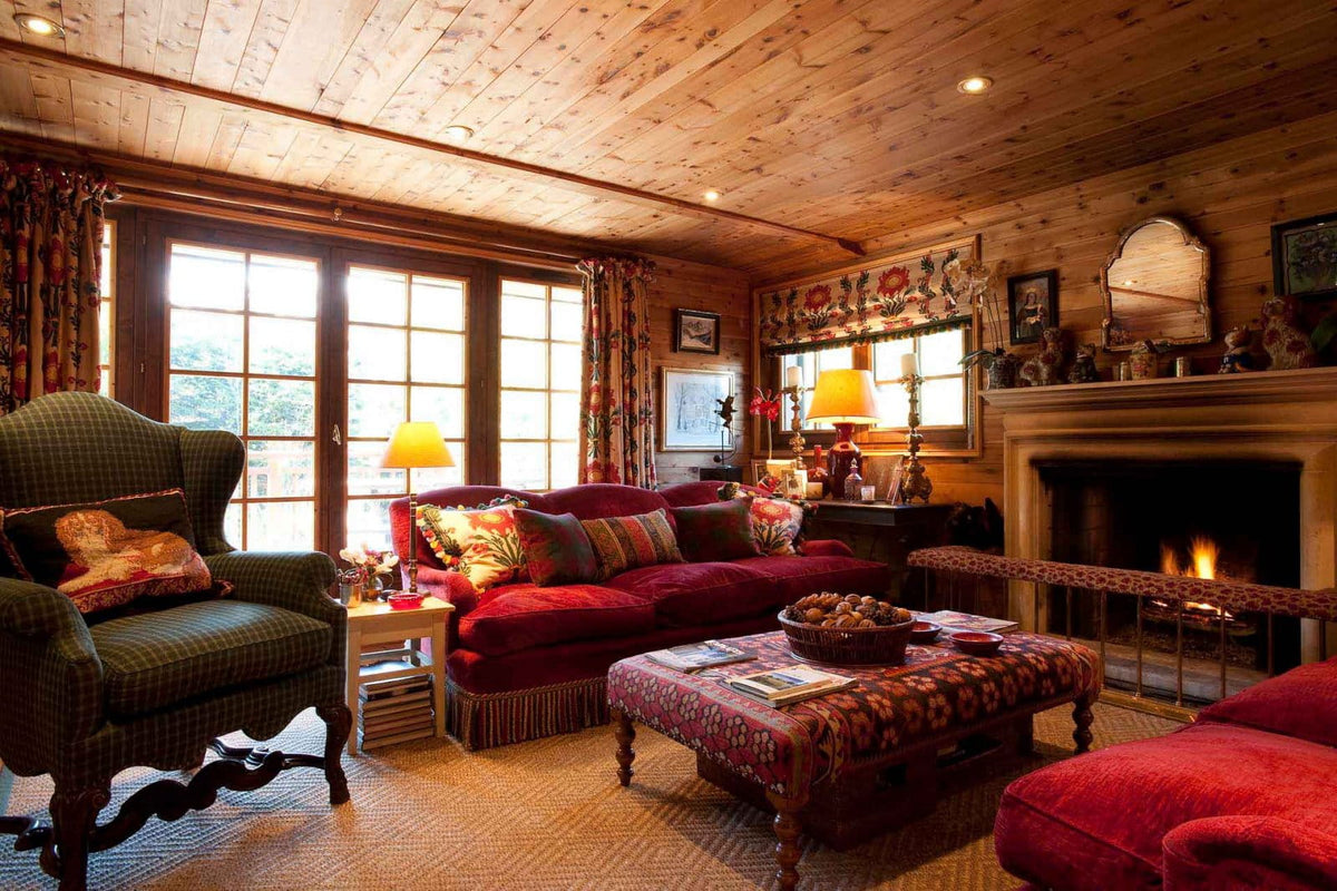 A cozy, rustic living room with wood-paneled walls and ceiling, featuring a red sofa, a green plaid wingback chair, a patterned coffee table, and a fireplace with a visible fire.