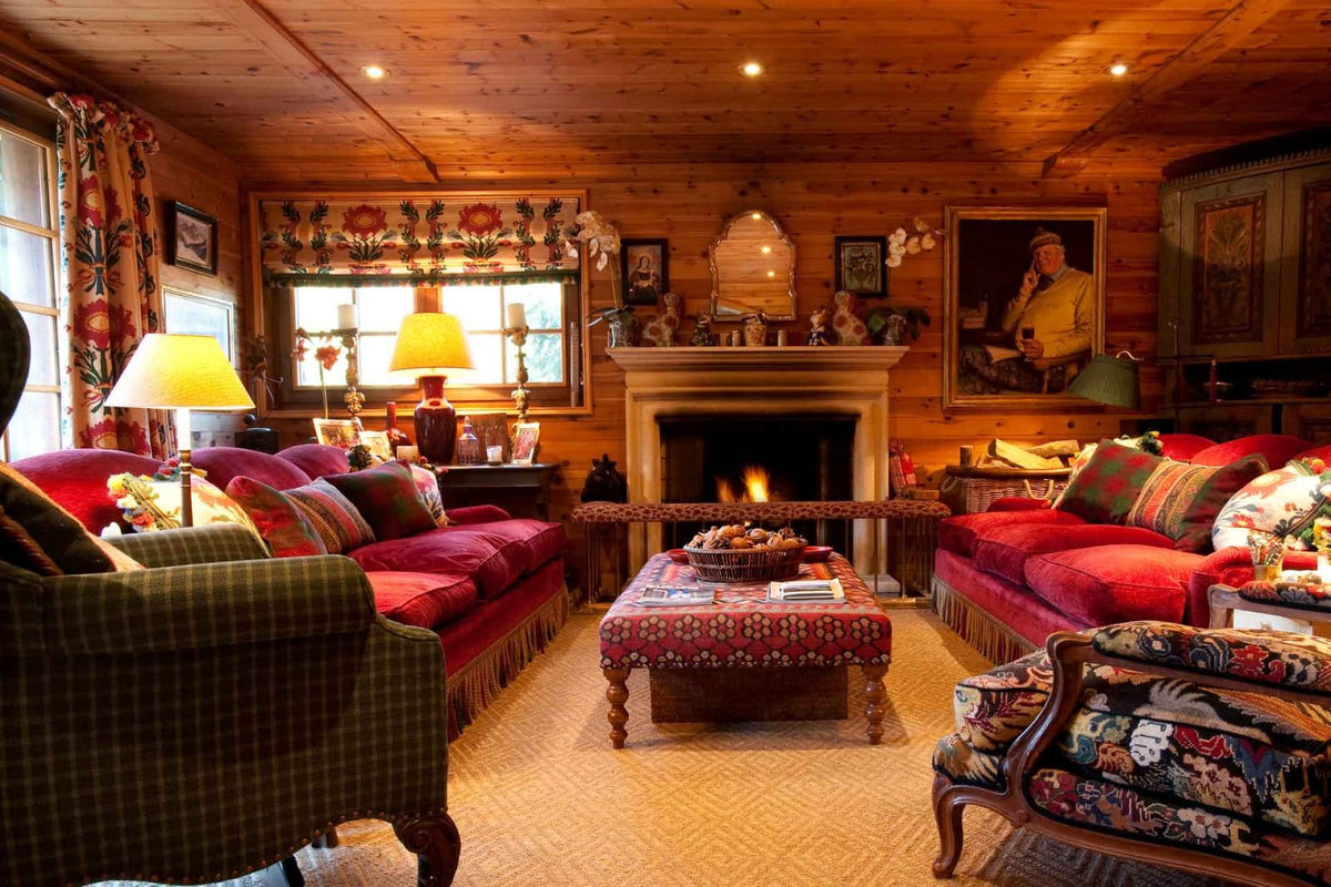 A cozy, rustic mountain-style living room with wood-paneled walls and ceiling, featuring red sofas, a green plaid armchair, a patterned coffee table, and a fireplace with a lit fire.