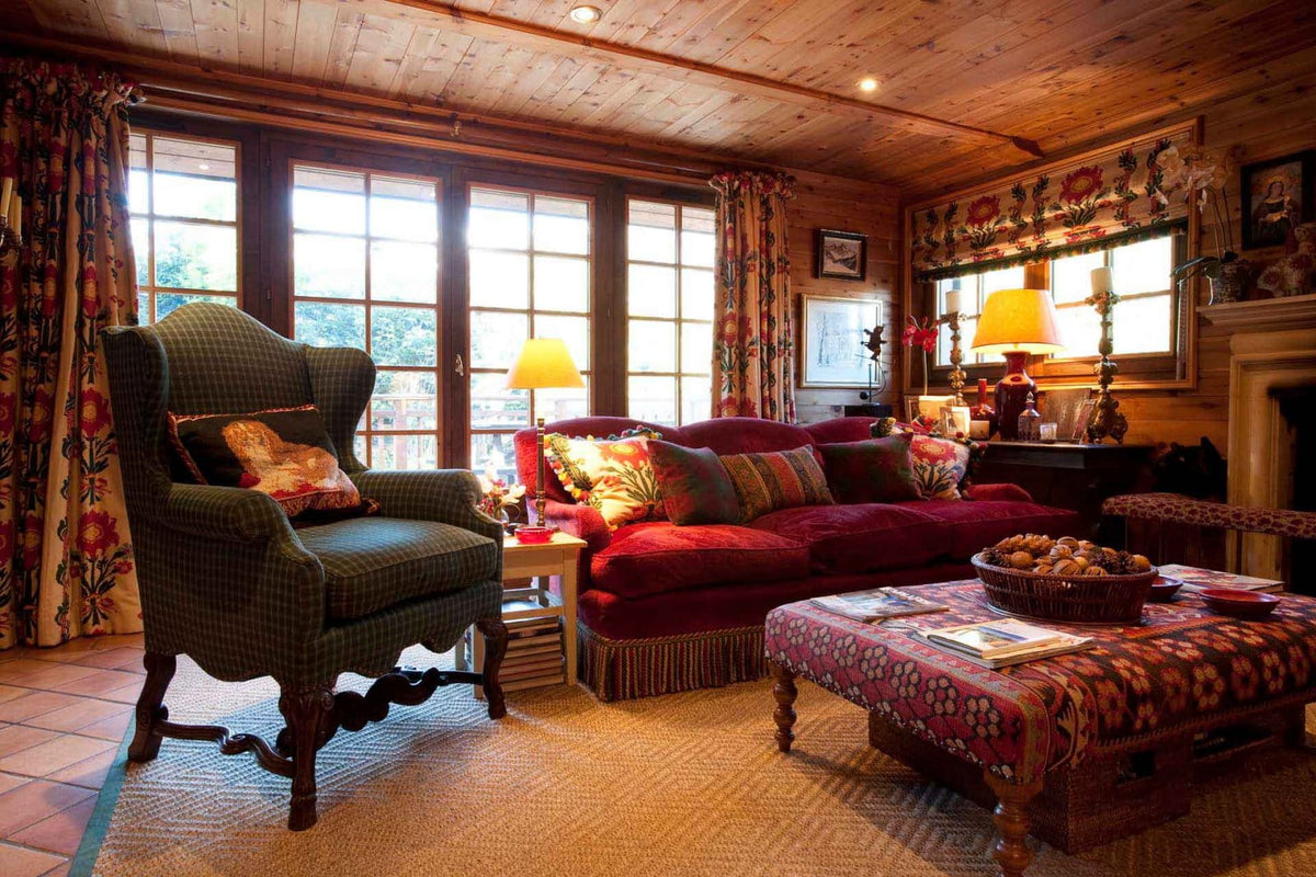 A cozy rustic living room with wood-paneled ceilings and walls, featuring a deep red velvet sofa with patterned pillows, a dark green plaid wingback armchair, a patterned coffee table, and large multi-paned windows with floral curtains.