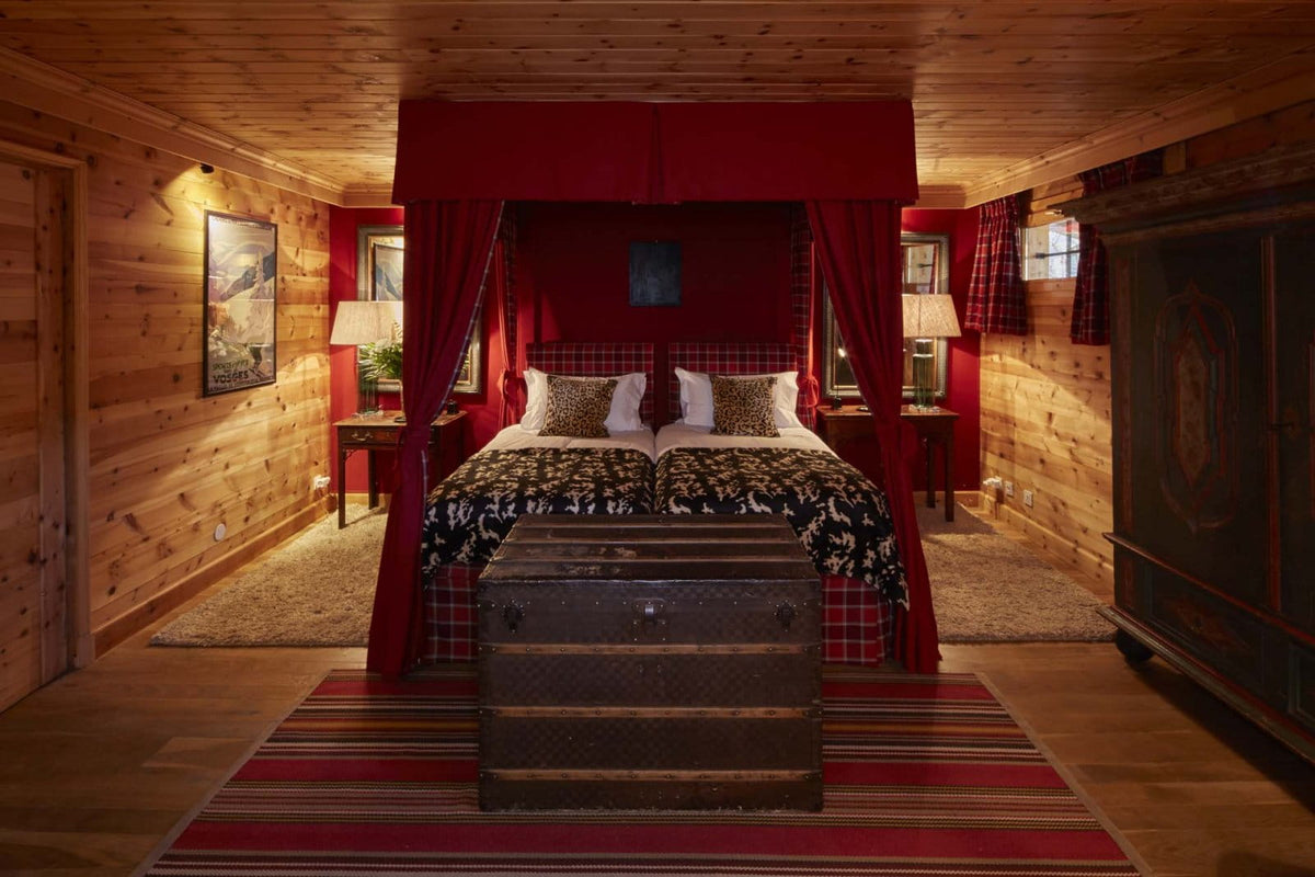 A rustic mountain chalet bedroom featuring a four-poster canopy bed with red drapes, a dark patterned comforter, a vintage-style trunk at the foot of the bed, and warm wood-paneled walls and ceiling.