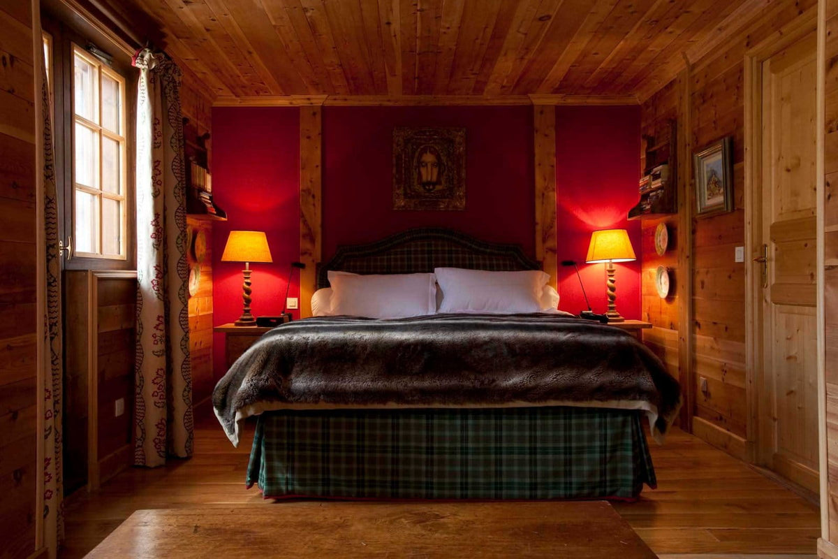 A rustic bedroom with a large bed dressed in dark gray and green plaid bedding, set against red walls with two lit table lamps on either side. The room has wood-paneled ceilings and walls, a window with curtains on the left, and a door on the right.