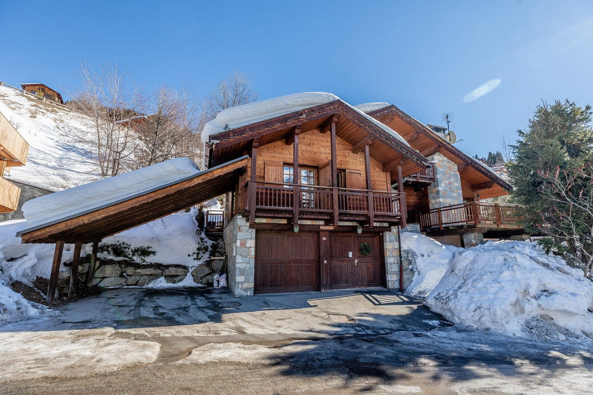 A large alpine chalet-style mountain house with snow-covered roofs and a covered driveway, set in a snowy outdoor setting under a clear blue sky.