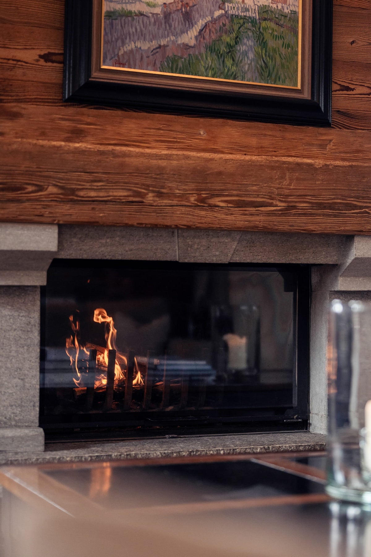 A cozy interior scene featuring a stone and wood fireplace with a visible fire, set within a rustic wooden wall. In the foreground, a glass candle holder and stacked books sit on a table, while a window in the background shows red cushions and a small figurine.