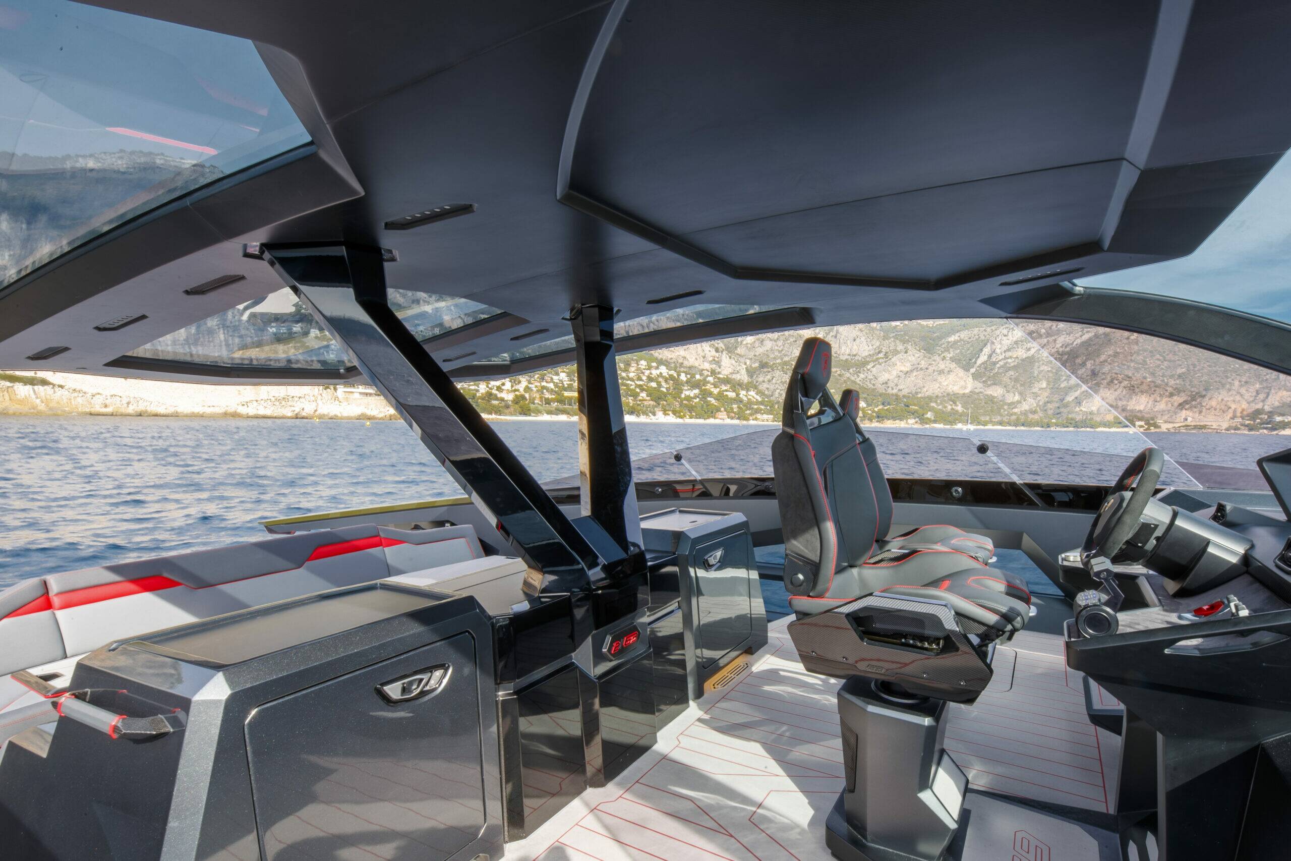 A luxury motorboat cockpit with a modern black and gray design, featuring a high-backed captain's seat, a console station, and a large wraparound windshield overlooking water and rocky mountains.