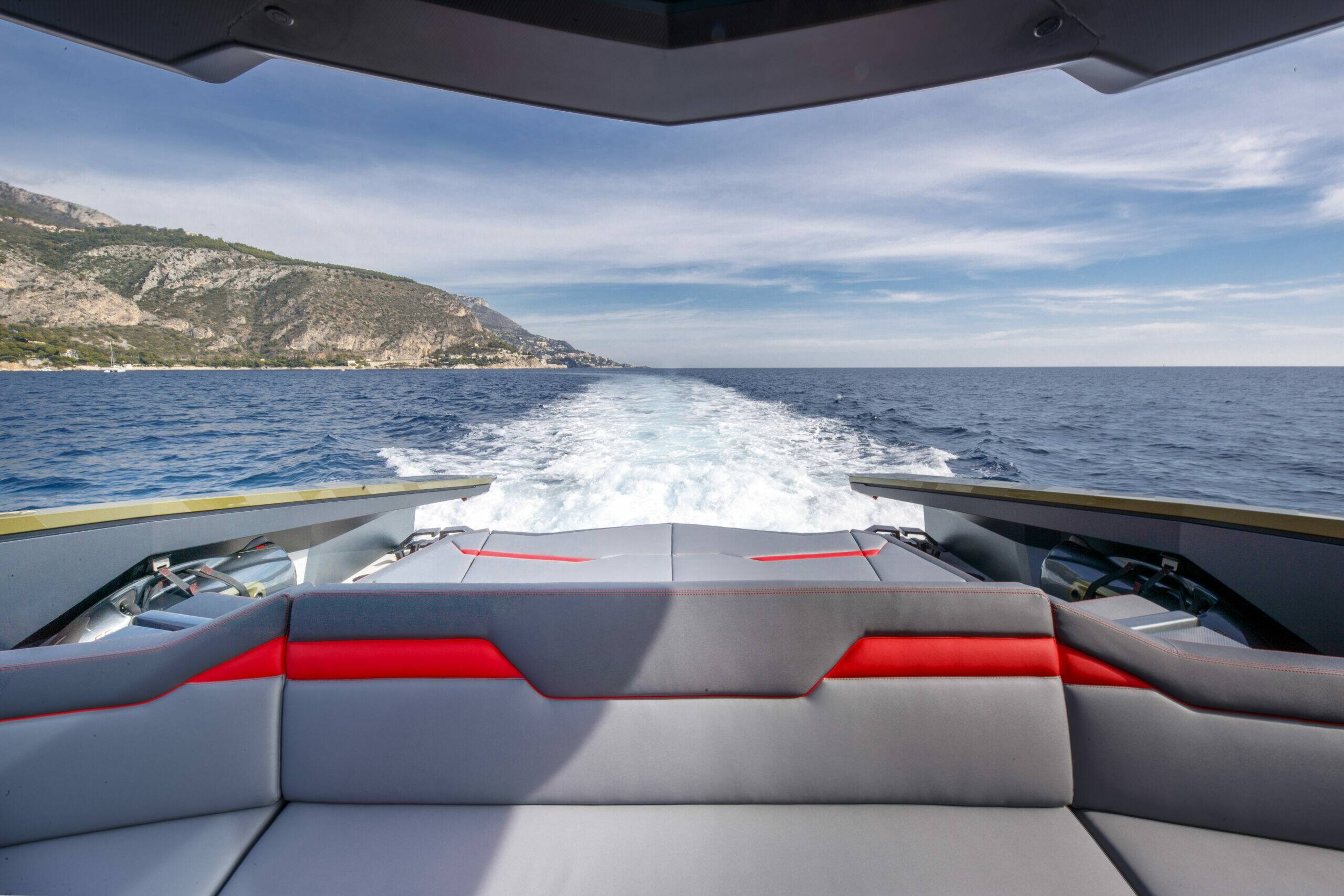 Rear view from inside a motorboat on open water, showing the wake behind the boat, a coastal shoreline in the distance, and a gray upholstered bench with red accent panels.