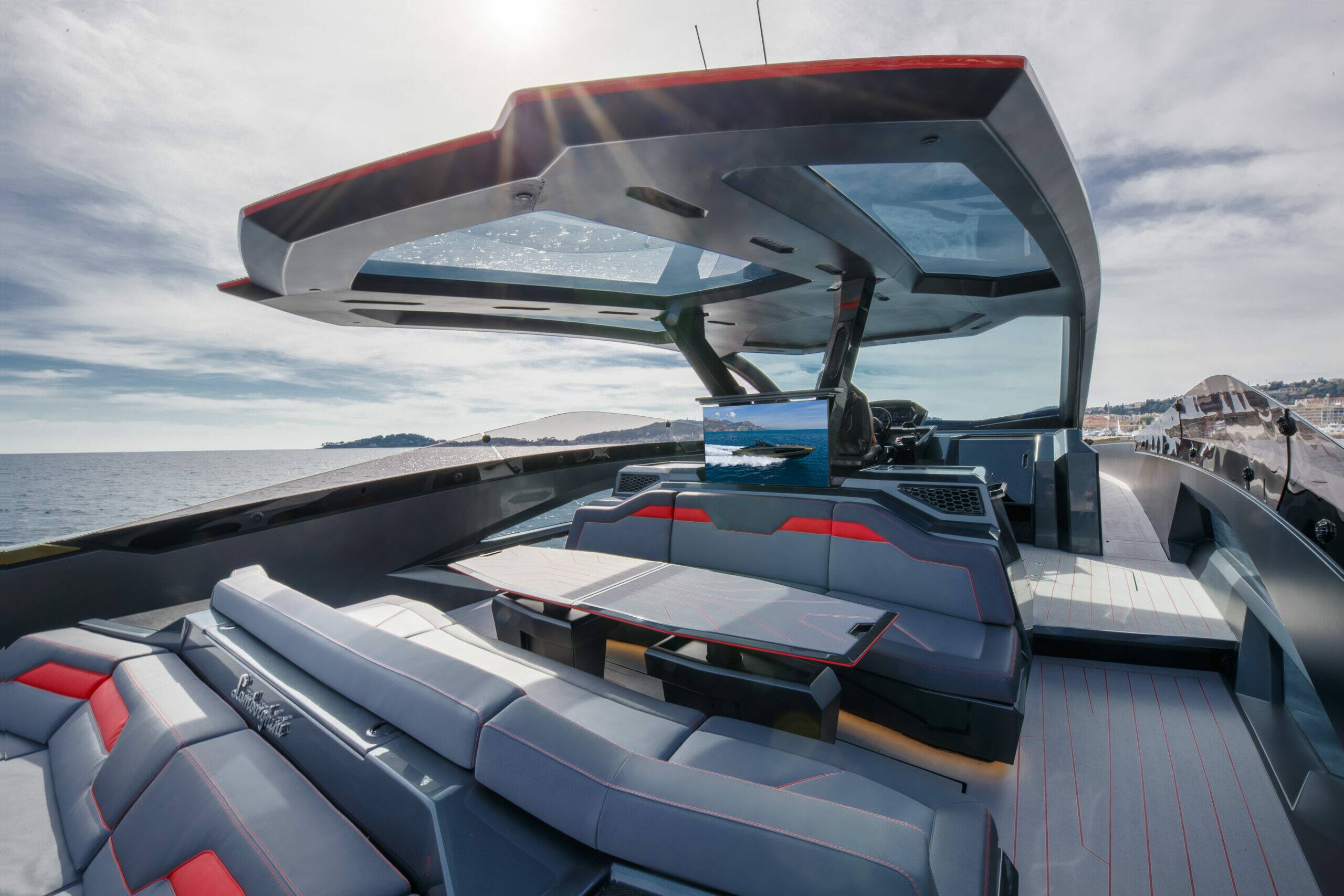 Luxury yacht cockpit and seating area featuring gray upholstered cushions with red stitching, a central table, a large curved windshield, and a sun canopy overlooking the open sea under a partly cloudy sky.