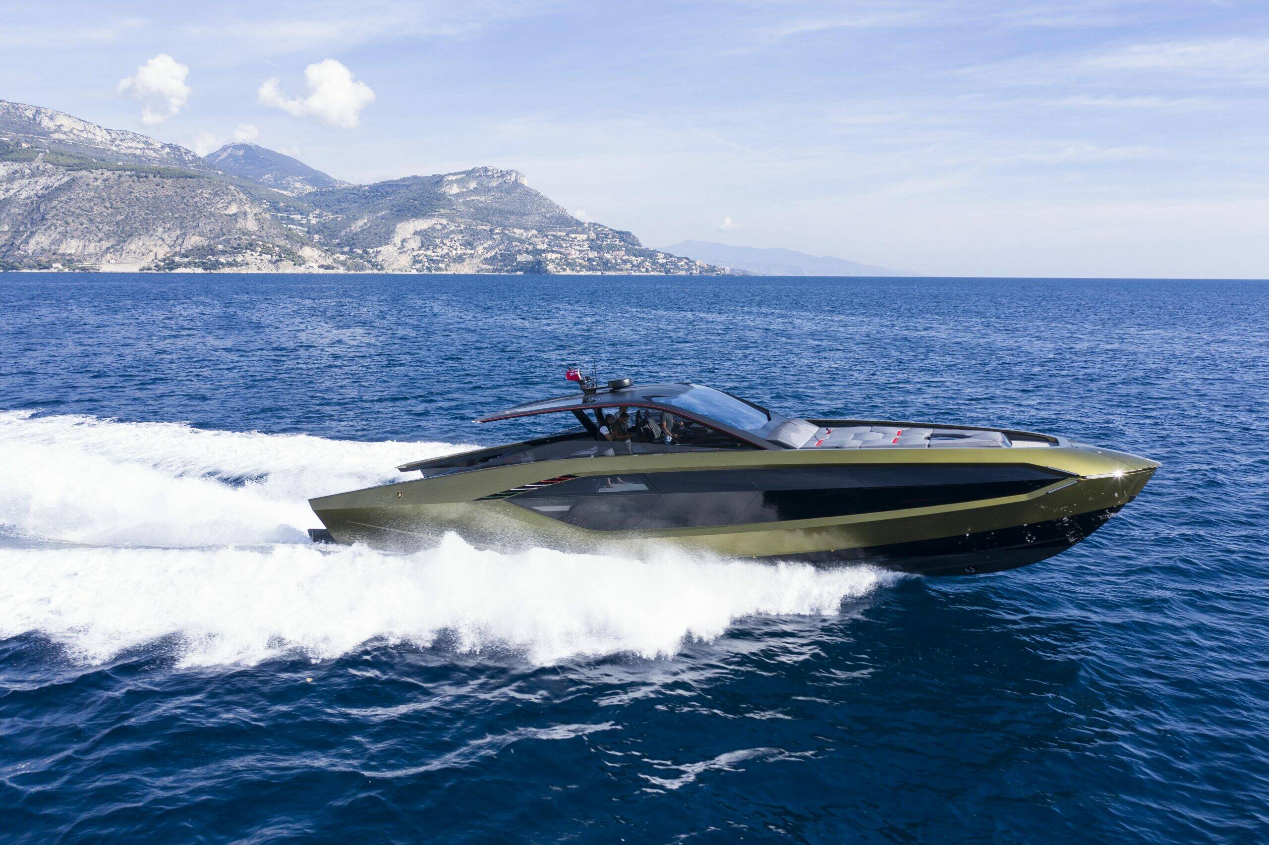 A sleek, high-performance speedboat in an olive green and black finish speeds across deep blue ocean water, leaving a white wake with mountains in the background under a partly cloudy sky.