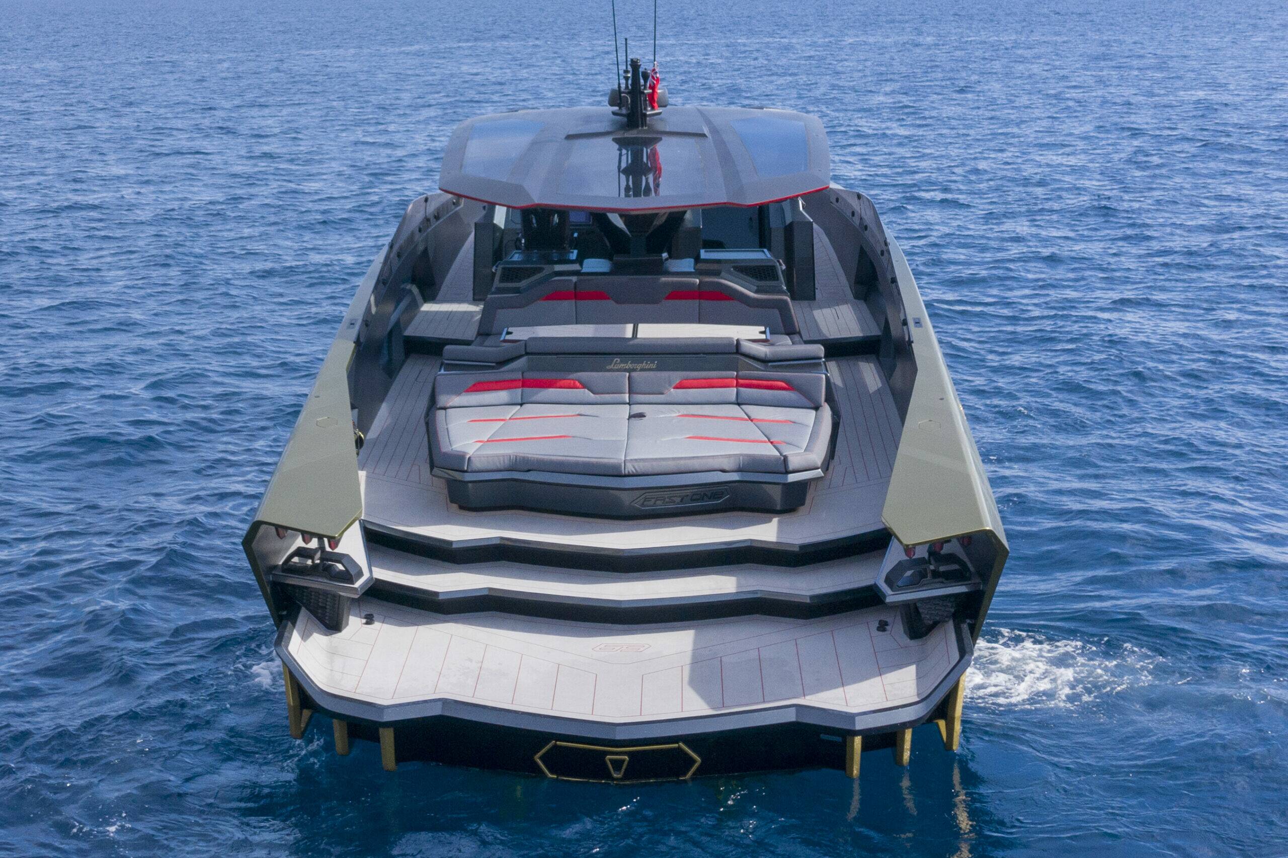 A luxury motor yacht shown from a high-angle rear view, sailing on deep blue ocean water. The yacht features a modern design with a gray and black exterior, a large sun deck with integrated seating, and a covered cockpit.