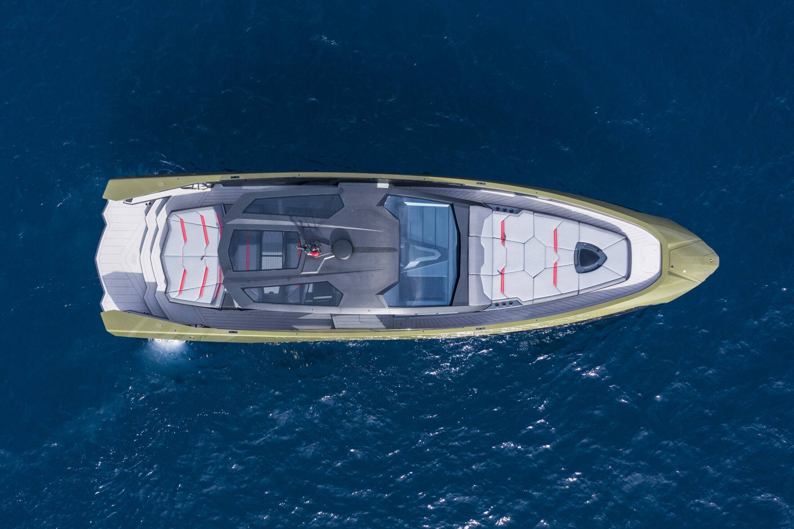A luxury motor yacht shown from a top-down aerial view, floating on deep blue ocean water. The boat features a metallic gold hull, a gray and white interior deck with red accents, and a transparent windshield.