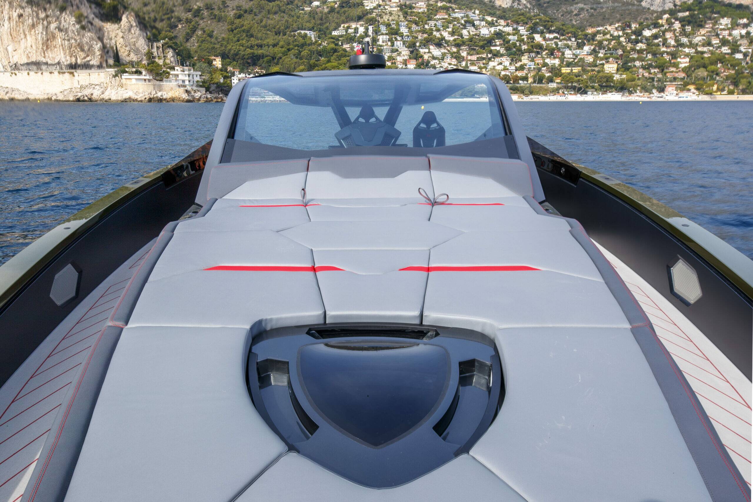 A close-up rear view of a modern luxury speedboat with a gray deck, red accent lines, and a dark windshield, sailing on blue water with a coastal hillside village in the background.