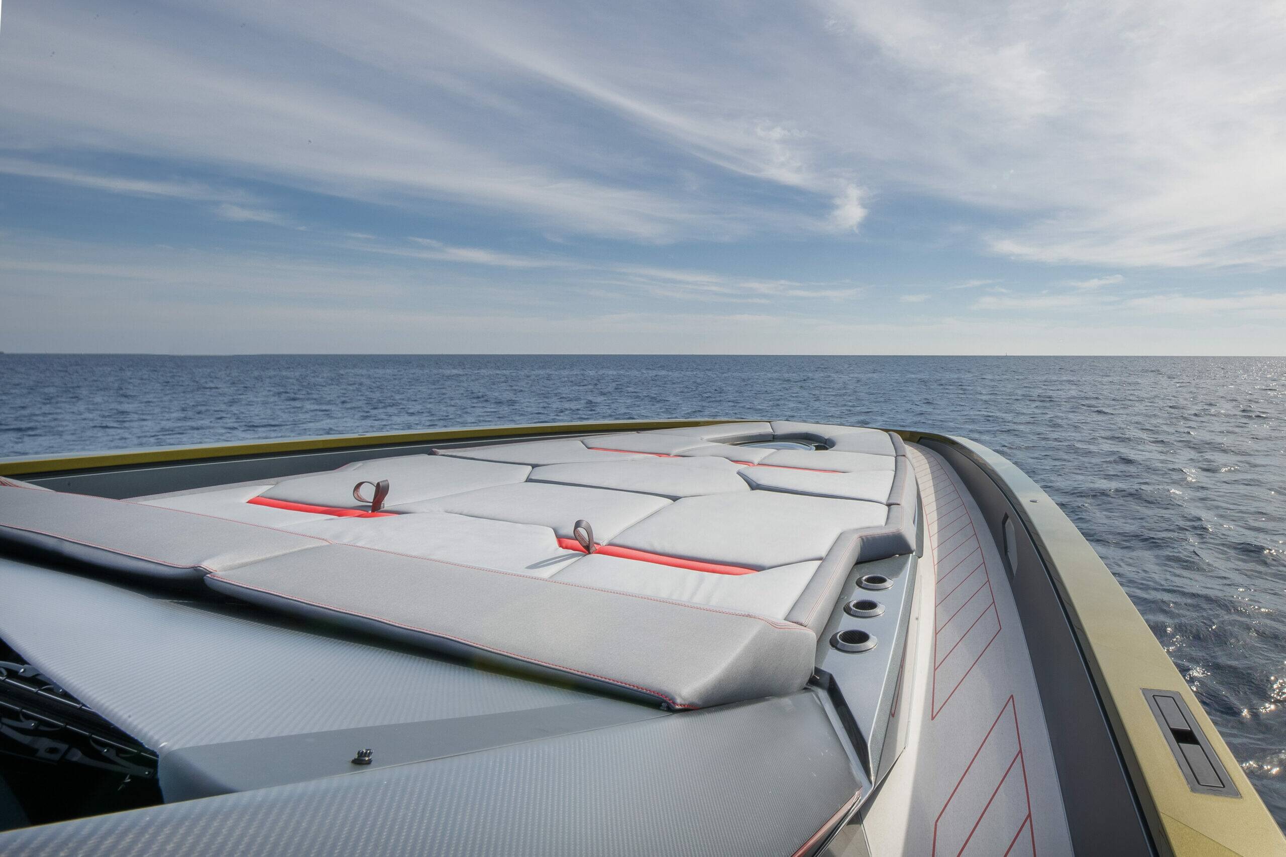 A close-up view of a boat deck with white cushioned seating and red trim, photographed from the bow while sailing on open blue water under a partly cloudy sky.