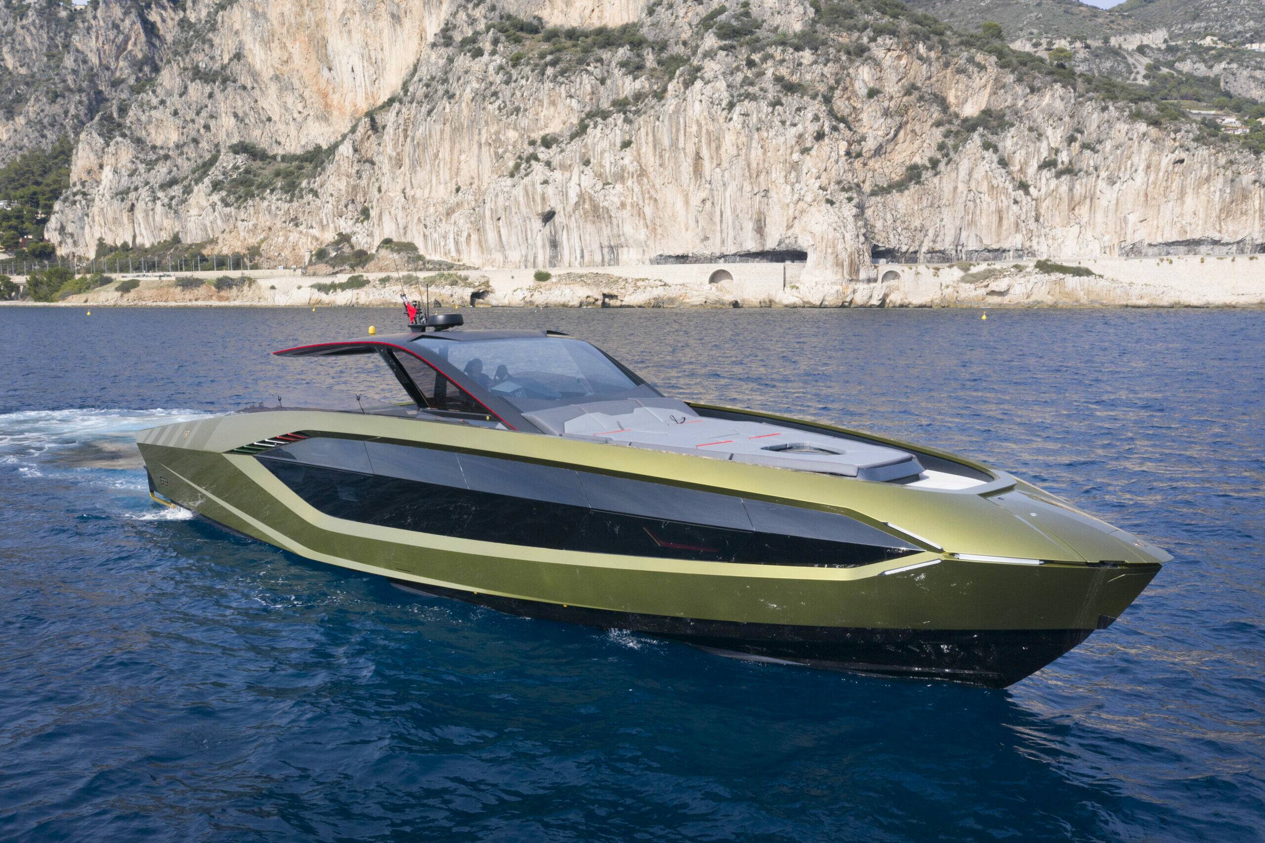 A modern speedboat with a metallic olive green hull and black accents, sailing on deep blue water with a rocky coastal cliff in the background.