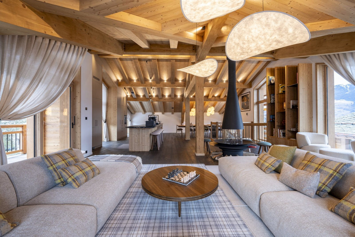 A luxurious chalet-style living room with light beige sectional sofas, plaid accent pillows, a round wooden coffee table, a black central fireplace, and a high-ceilinged rustic wooden ceiling with exposed beams.