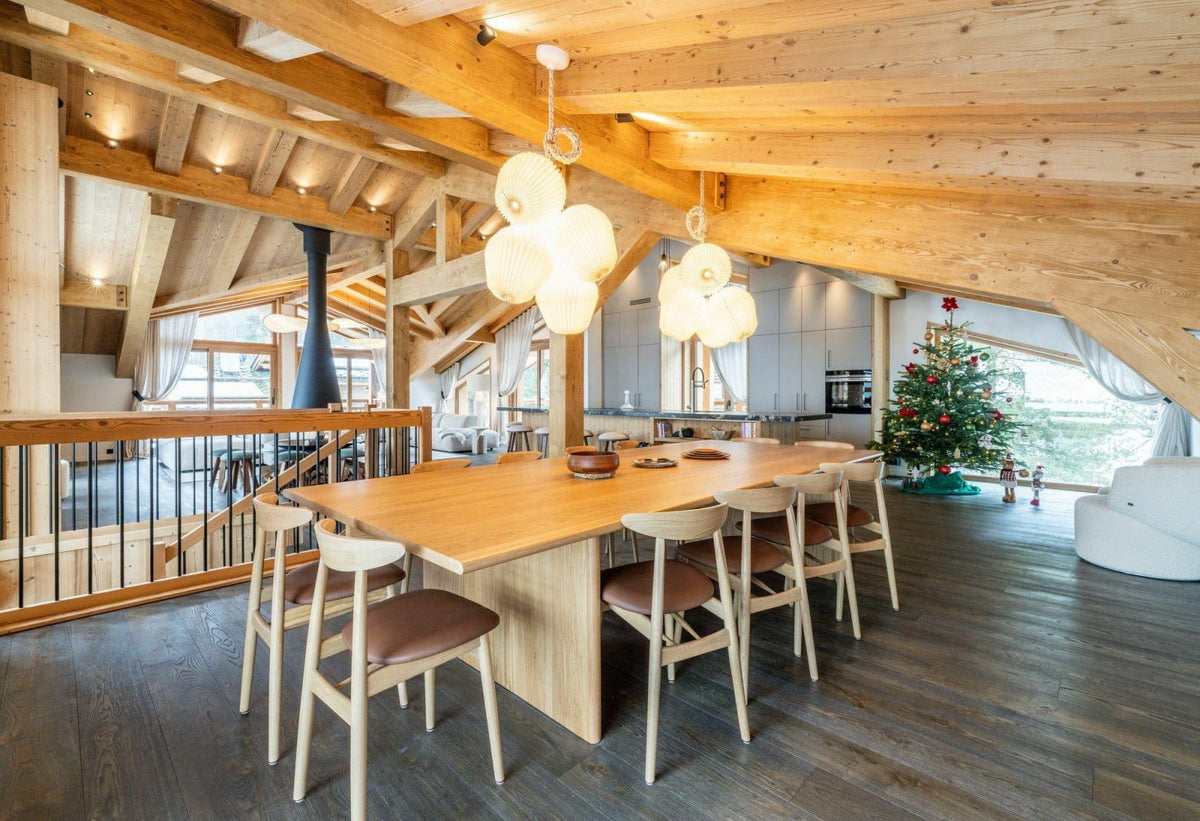 A modern wooden dining room with a long light oak table, six matching chairs with brown upholstered seats, and two sculptural white pendant lights hanging from a sloped wooden ceiling. A staircase, kitchen, and a decorated Christmas tree are visible in the open loft-style interior.