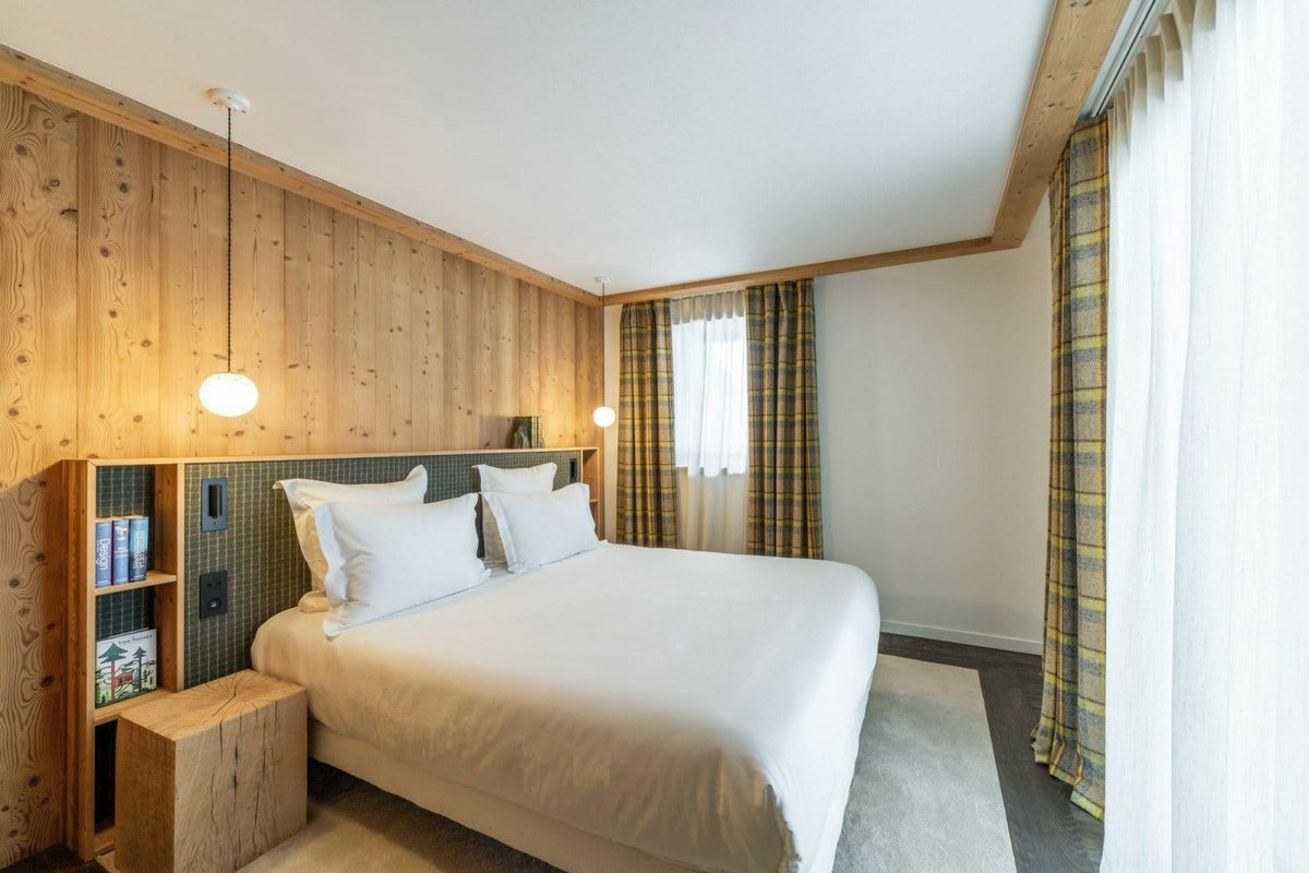A modern bedroom featuring a neatly made bed with white linens, a light wood headboard and bedside table, wood-paneled walls, plaid curtains, and a bright window.