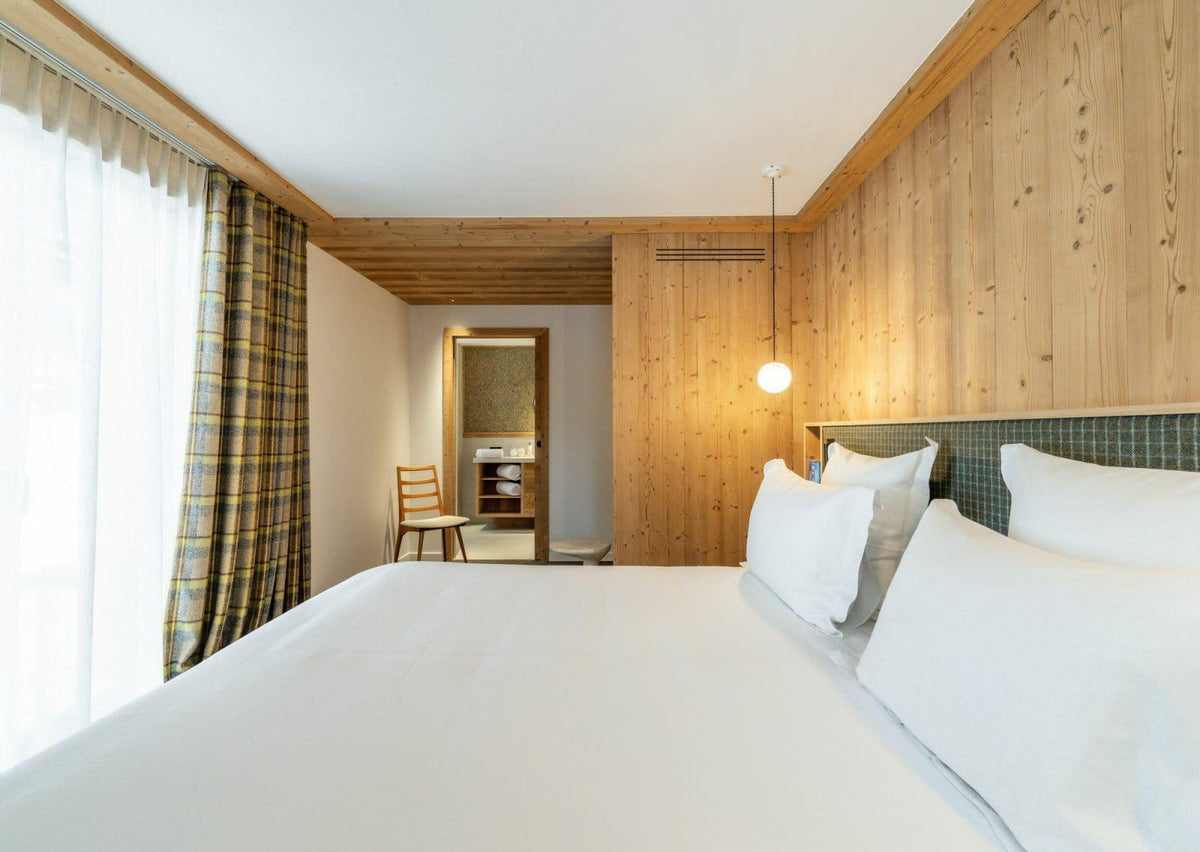 A modern bedroom featuring a large bed with white bedding, a wood-paneled wall, and plaid curtains by a bright window.