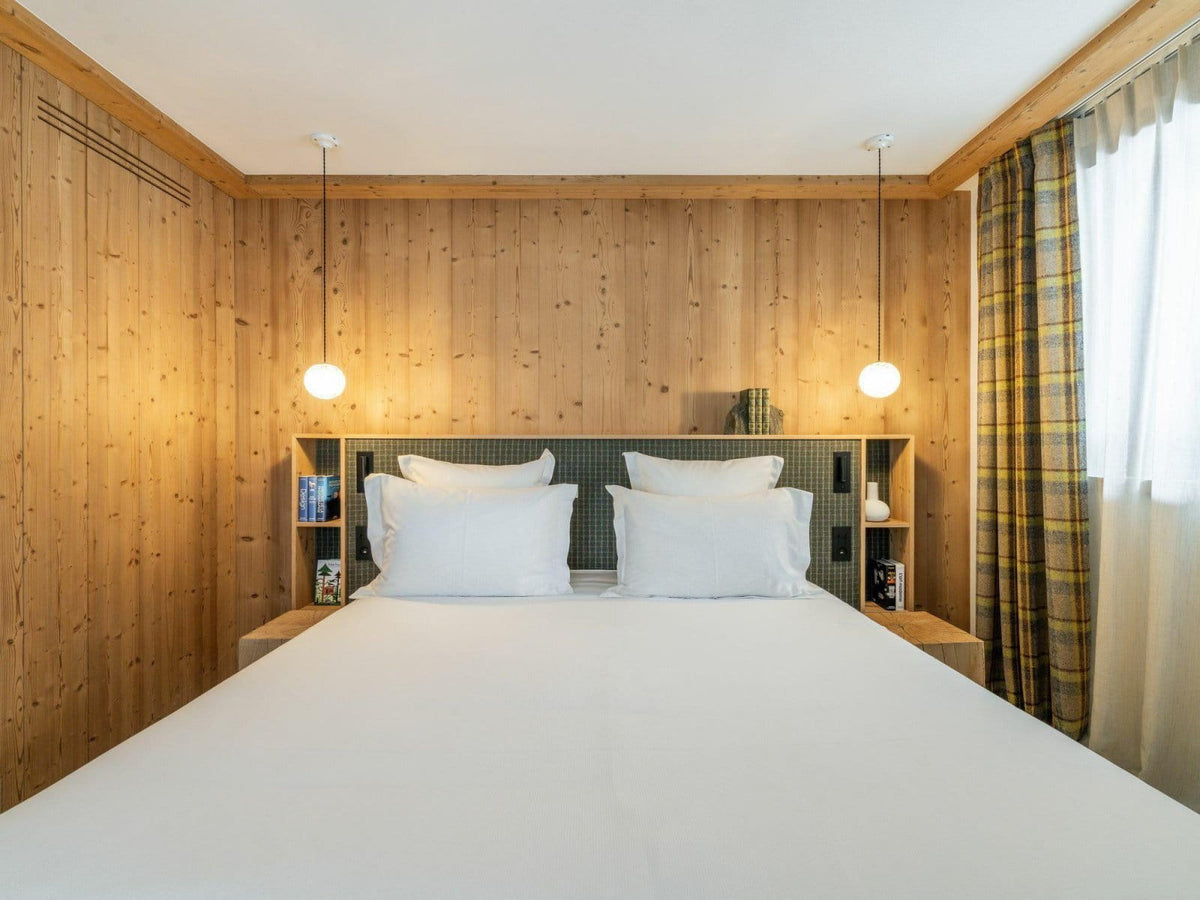 A cozy bedroom with a neatly made bed featuring white linens and pillows, set against a light wood-paneled wall with built-in shelving and two hanging pendant lights.