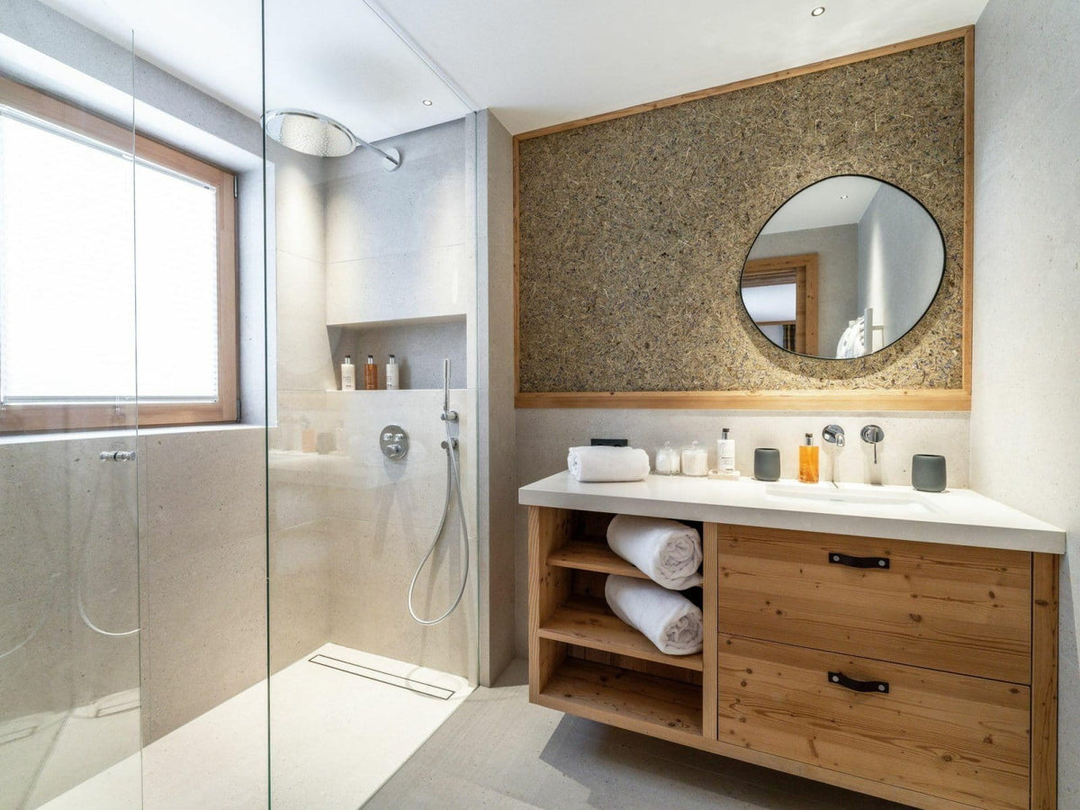 Modern bathroom vanity with a light wood cabinet, white countertop, round mirror, and textured wall panel, shown in a bright bathroom with a glass shower enclosure.
