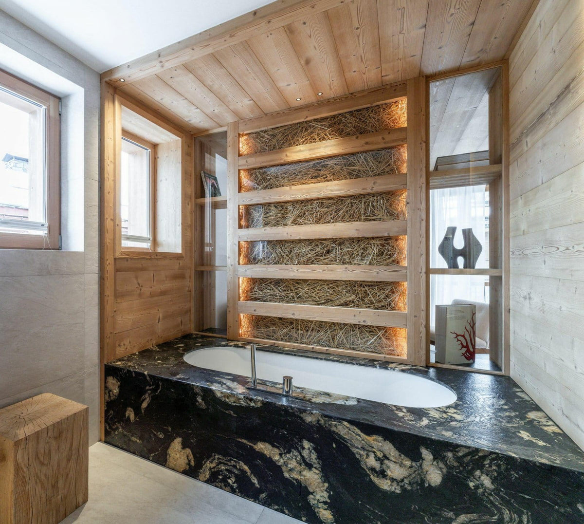 A luxury modern bathroom featuring a built-in oval bathtub set into a black and gold marble base, surrounded by light wood paneling and a decorative straw wall with warm lighting.