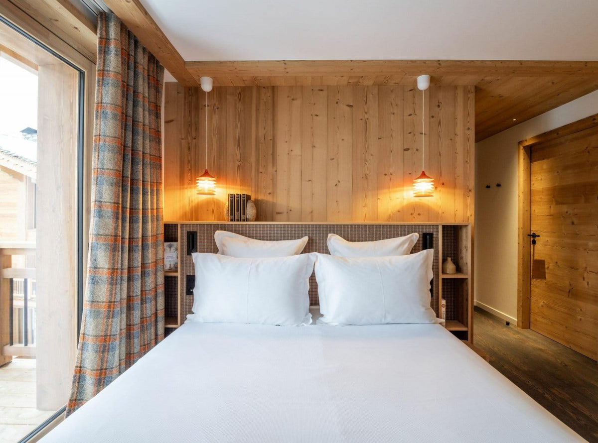 A modern bedroom interior with a neatly made bed in white linens, set against a wood-paneled wall with two lit pendant lamps. A plaid curtain hangs to the left, and a wooden door is visible on the right.
