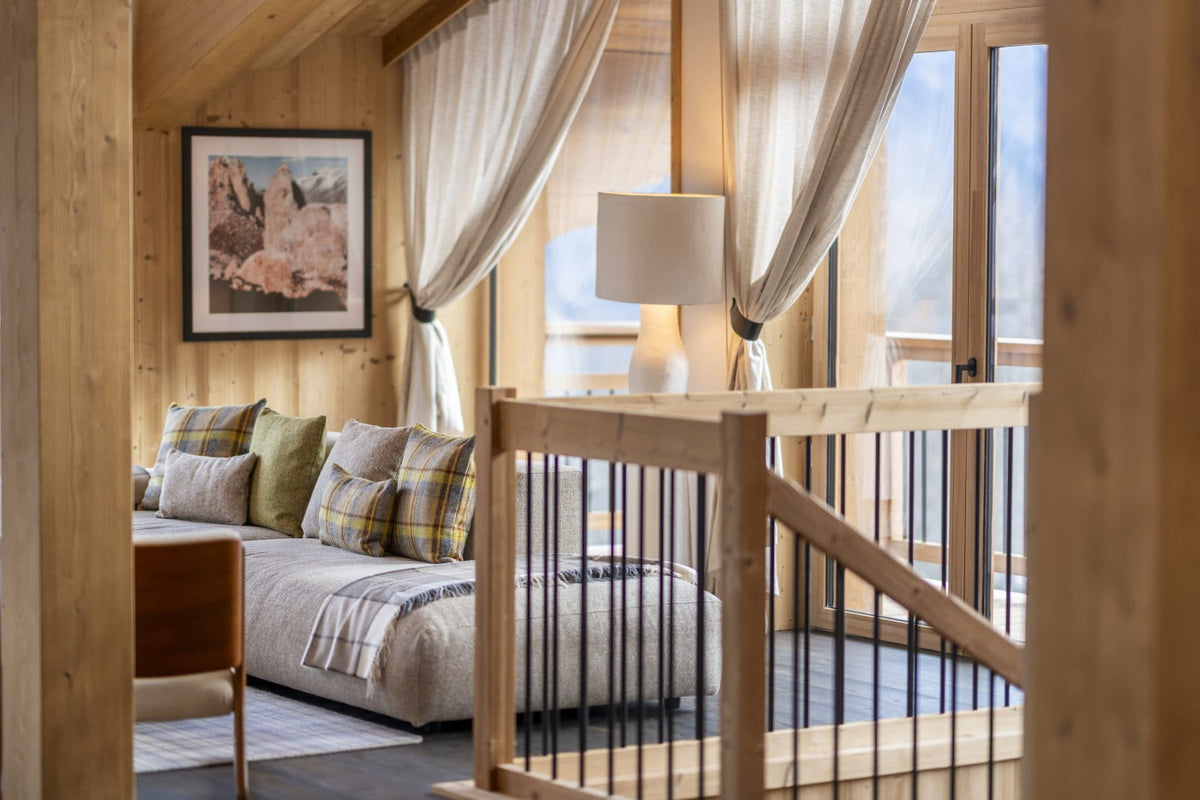 A cozy mountain chalet bedroom featuring a bed with plaid accent pillows, a wooden staircase railing in the foreground, and large windows with sheer curtains looking out to a snowy mountain view.