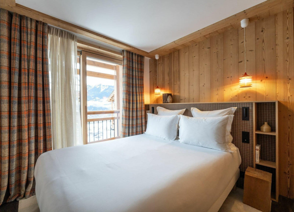 A cozy mountain-view bedroom with a neatly made white bed, wood-paneled walls, plaid curtains, and warm bedside lighting.