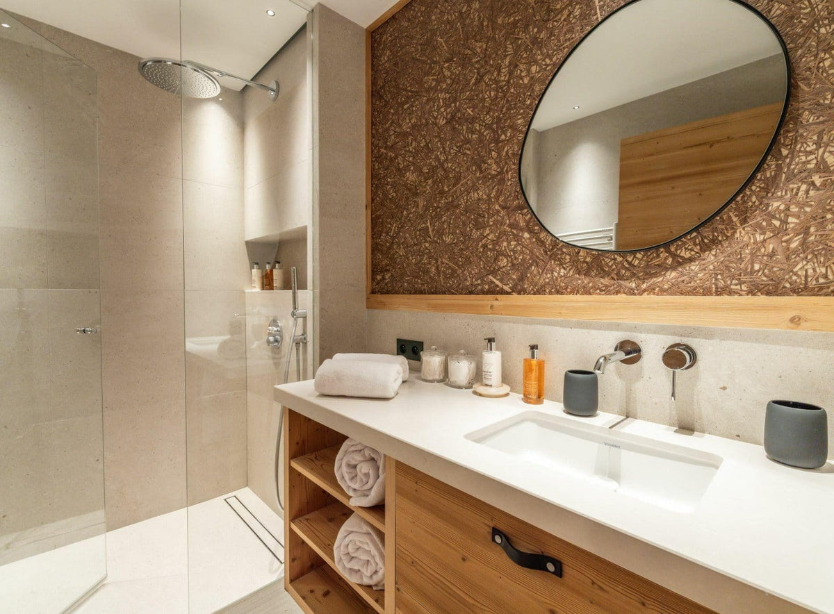 Modern bathroom vanity with a white countertop, open wood cabinet, oval mirror, and a glass-enclosed shower area.