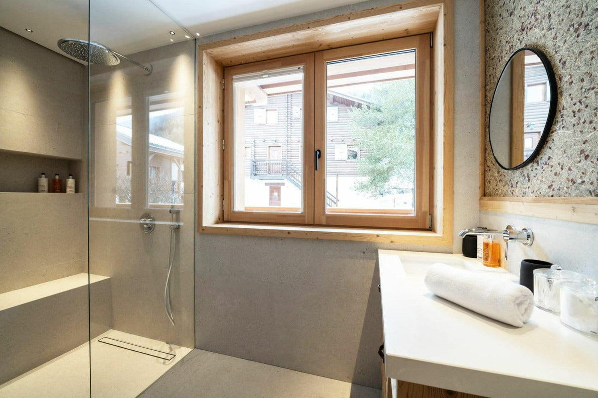Modern minimalist bathroom interior featuring a glass shower enclosure, a large wood-framed window looking out onto buildings, a white vanity with a rolled white towel, and a round black-framed mirror.