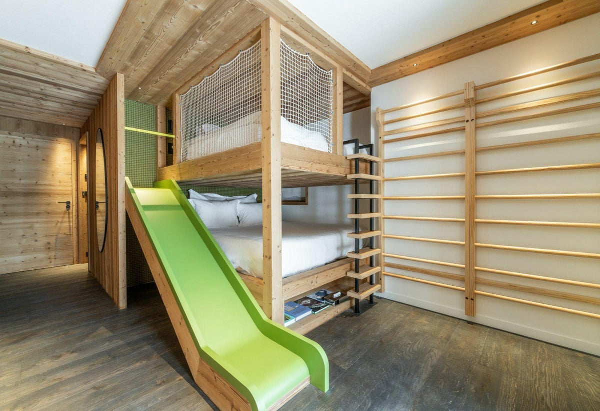 A rustic wooden bunk bed with a bright green slide and a ladder, set in a wood-paneled room with wall gymnastics bars.