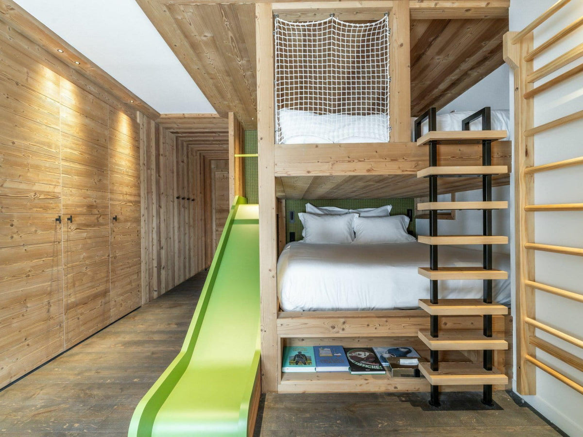 A compact wooden bunk bed with a bright green slide, a small ladder, a net top, and white bedding, set against a wood-paneled wall.