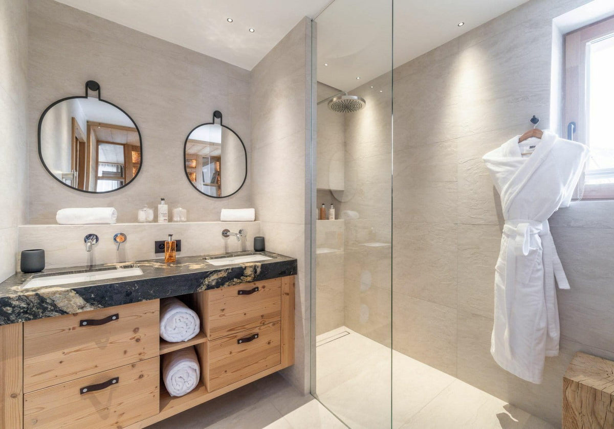 Modern bathroom interior with a double-sink vanity made of light wood and black marble, two round mirrors, a glass shower enclosure, and a white bathrobe hanging on a wall hook.
