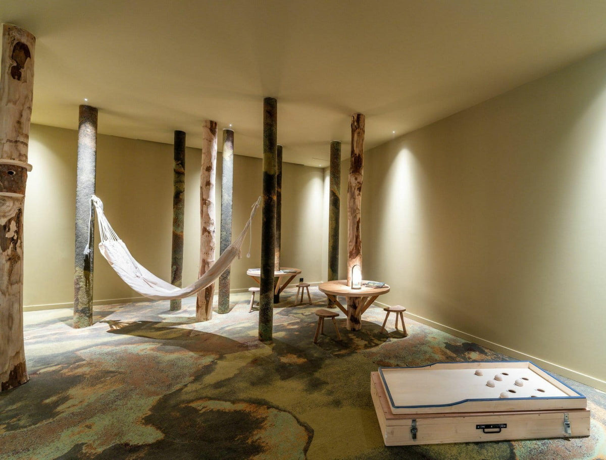 A rustic indoor lounge space with a white fabric hammock suspended between tall natural wood posts, a small round wooden table with stools, a patterned carpet, and a game board case in the foreground.
