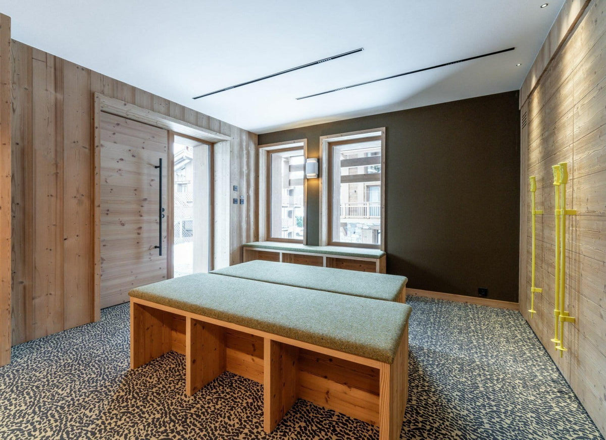 A modern interior room with light wood paneled walls, a dark brown accent wall, a patterned floor, and two low wooden benches with light green upholstered tops placed in the foreground.