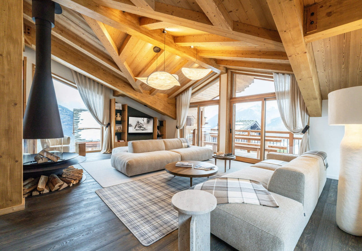 A modern chalet-style living room with a light beige sectional sofa, a round coffee table, a plaid rug, a black freestanding fireplace with visible logs, large wood-beam ceilings, and floor-to-ceiling windows overlooking a mountain view.