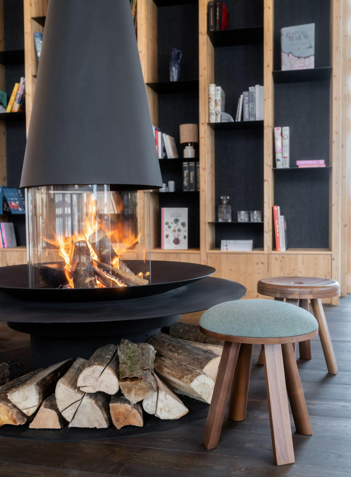 A modern black metal fireplace with a tall conical hood and visible flames, set in a cozy room with wood shelving, stacked firewood, and two wooden stools.