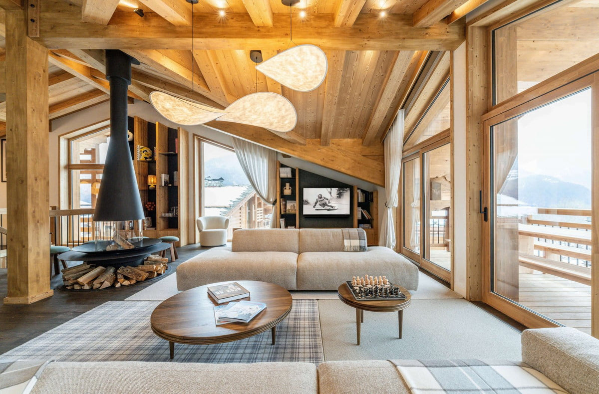 A luxury modern chalet living room with a light beige sectional sofa, round wooden coffee tables, a black conical fireplace chimney, and large glass windows looking out to a mountain view.