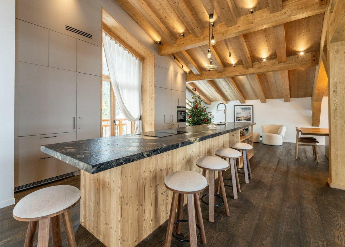 A modern kitchen island with a light wood base, a dark stone countertop, and six matching bar stools, set in a rustic room with exposed wooden ceiling beams and dark wood flooring.