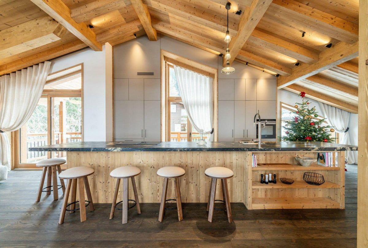 A modern alpine-style kitchen island with a light wood base, dark stone countertop, four wooden bar stools, and a decorated Christmas tree in the background under a sloped wooden ceiling.