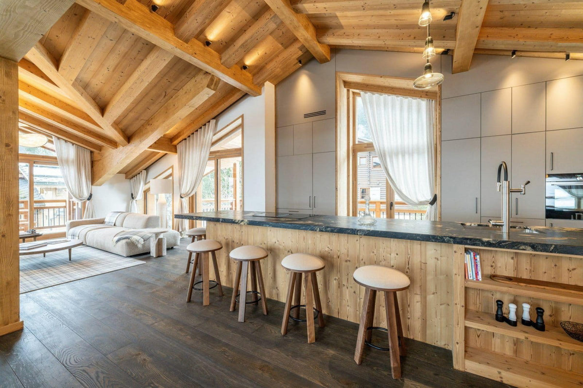A modern ski chalet kitchen and bar area with light wood cabinetry, a dark stone countertop, four bar stools, exposed wooden ceiling beams, and large windows with white curtains.