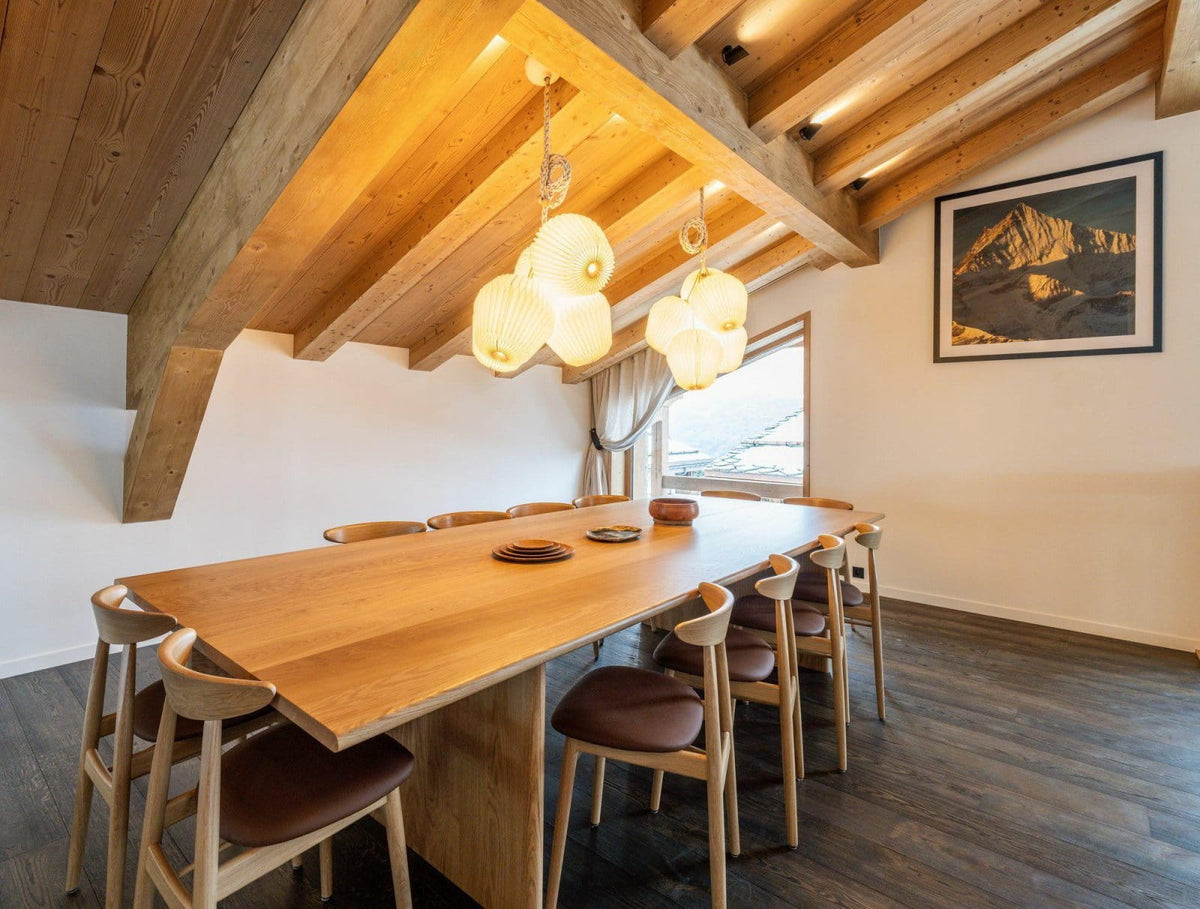 A modern dining room with a long wooden table and matching wooden chairs, under a sloped wooden ceiling with exposed beams and hanging pendant lights. A framed landscape print hangs on the wall.