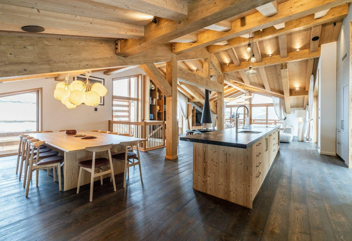 A luxury modern chalet kitchen and dining area featuring a large wooden island with a dark countertop, a dining table with wooden chairs, exposed wooden ceiling beams, and dark wood floors.
