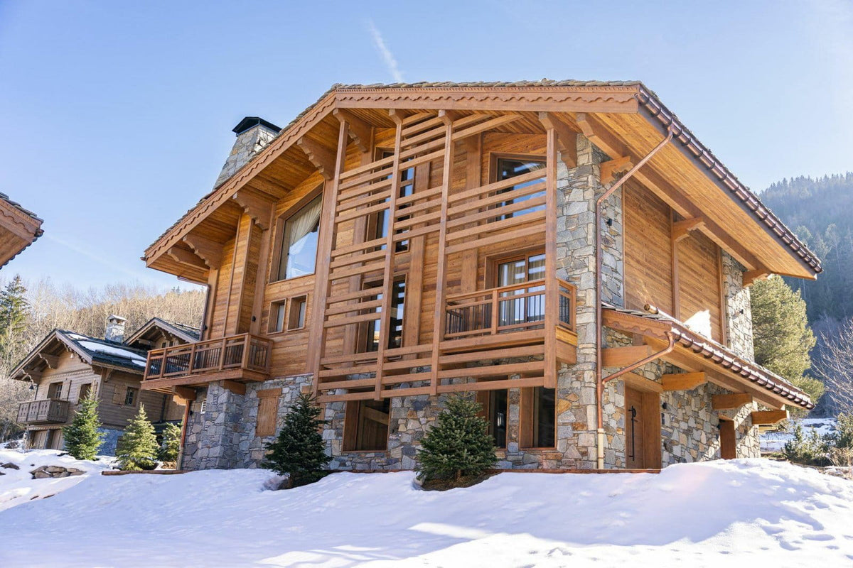 A large alpine-style wooden chalet with stone foundations, snow-covered ground, trees, and mountains in the background under a clear blue sky.