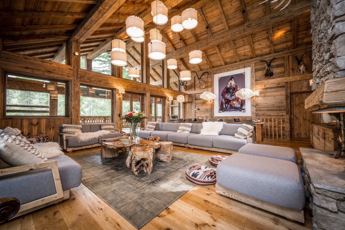 A luxury chalet-style living room featuring light gray upholstered sofas, a rustic wood log coffee table, a large gray area rug, a stone fireplace, and a high wooden ceiling with multiple hanging pendant lights.