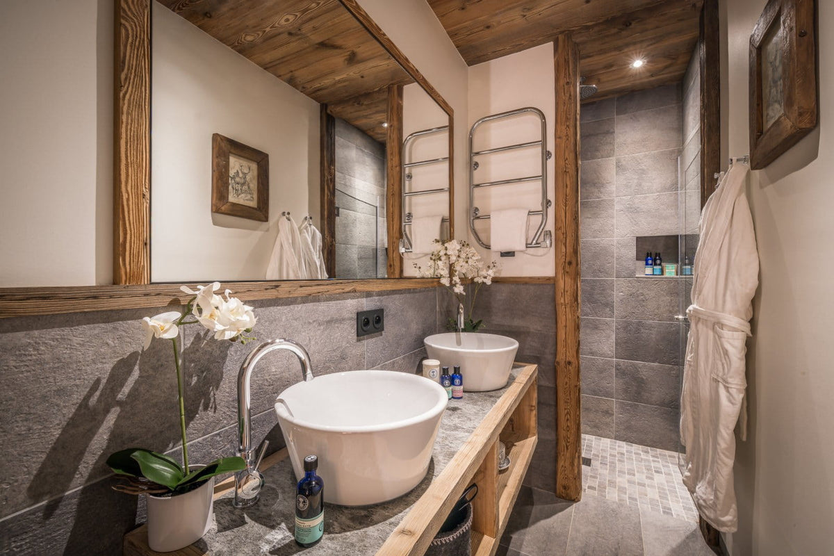 A modern rustic bathroom featuring two white vessel sinks on a wooden vanity, a large mirror, gray stone walls, a walk-in shower, and a white robe hanging on the right.