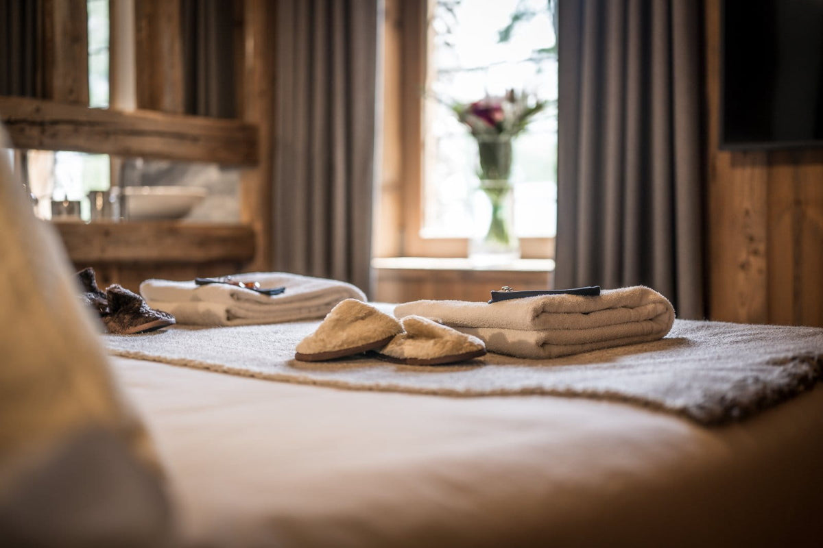 A cozy bedroom scene with a neatly made bed in the foreground, folded beige towels, a pair of slippers, and a dark remote on a textured rug. A softly blurred window, curtains, and wooden wall paneling are visible in the background.