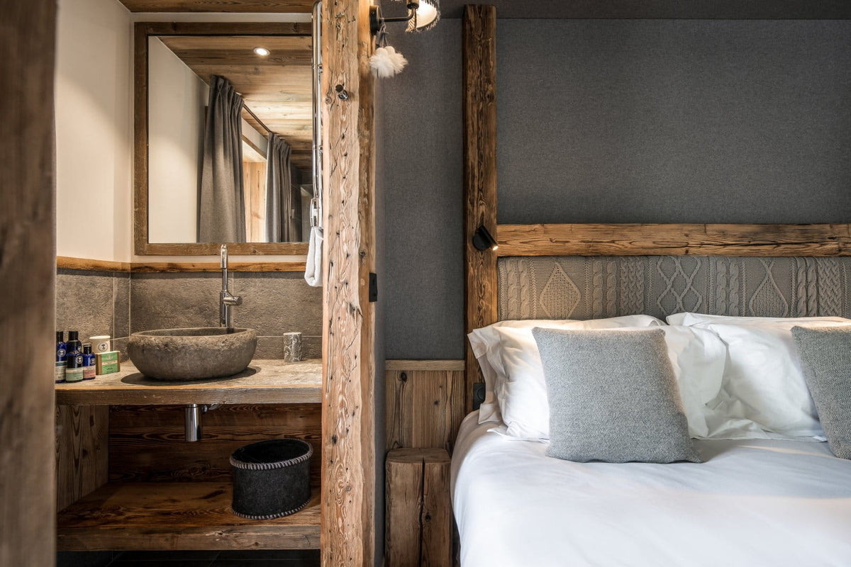 A rustic bedroom interior with a large bed dressed in white linens and gray pillows, set beside an open doorway revealing a matching wood vanity with a stone sink and mirror.