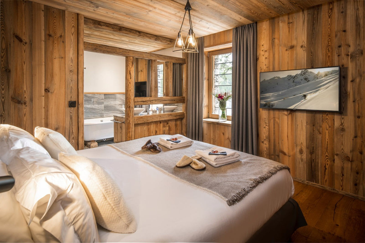 A cozy bedroom with rustic wood-paneled walls and ceiling, featuring a neatly made bed with white linens and a gray throw blanket. A wall-mounted landscape print, a window with curtains, and a mirror are visible in the room.