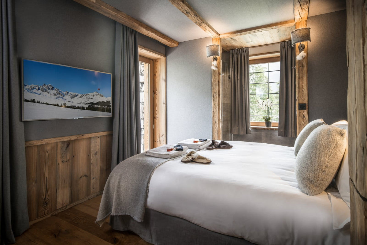 A modern rustic bedroom featuring a large bed with white linens and textured beige pillows, set against dark gray walls with a wooden wall panel and a framed mountain landscape print. The room has exposed wooden beams, a window with gray curtains, and warm ambient lighting.