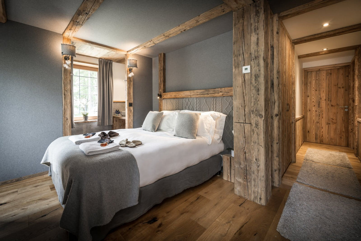 A rustic bedroom interior with a neatly made bed dressed in white linens and a gray throw, set in a cabin-like room with exposed wooden beams, wood flooring, and a wood-paneled hallway.