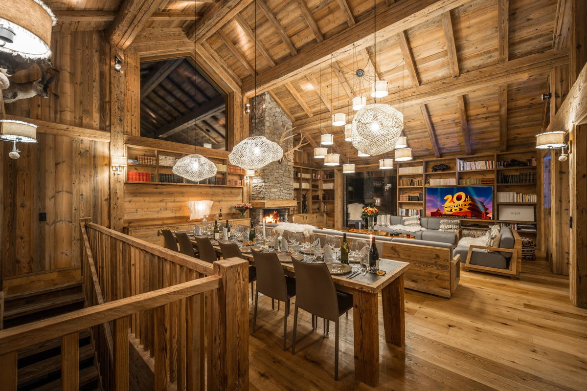 A luxurious alpine chalet interior featuring a rustic wooden dining room with a long wooden dining table, chairs, and pendant lights, connected to a modern living room with a fireplace and TV.